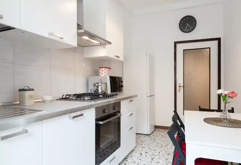 Cucina moderna e luminosa con pensili bianchi, piano cottura a gas, forno incassato e piccolo tavolo da pranzo. Superfici pulite, contenitori decorativi e un vaso di fiori rendono l'ambiente accogliente.