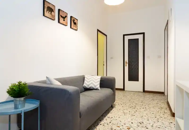 Soggiorno compatto e ben illuminato con un divano grigio con cuscini, un piccolo tavolino blu con pianta in vaso e semplici quadri a parete — pulito, moderno e accogliente.