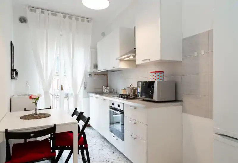 Cucina moderna e luminosa con mobili bianchi, forno incassato, piano cottura a gas e un piccolo tavolo da pranzo vicino alla finestra che dà sul balcone. Dettagli curati come i cuscini rossi e un vaso rendono l'ambiente accogliente per un appartamento compatto.