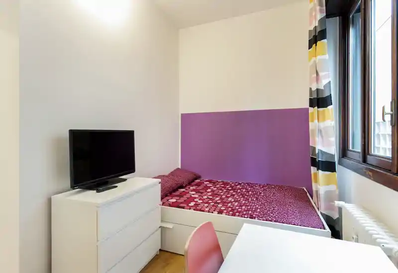 Compact bedroom with a single bed dressed in a patterned red duvet, a white chest of drawers with a TV, desk and a large window providing natural light.