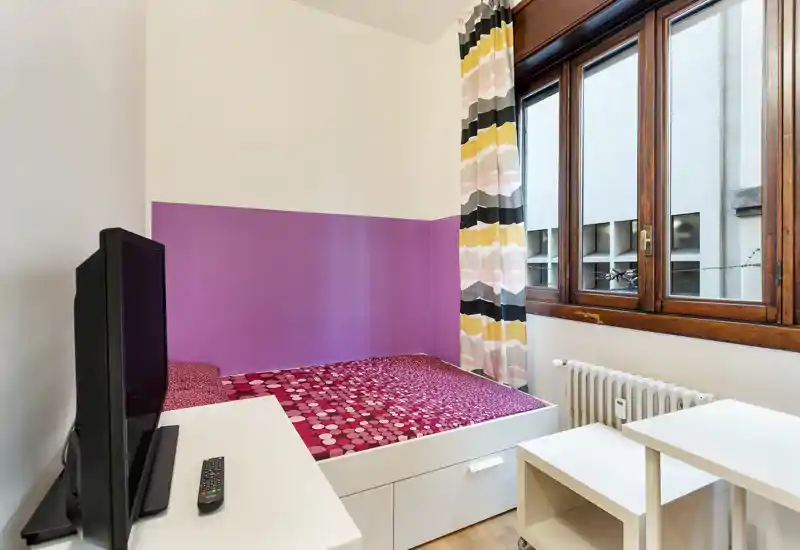 Small furnished bedroom with a single bed featuring patterned bedding, a large wooden-framed window providing natural light, a radiator and compact white storage furniture—bright, colorful and neatly arranged.