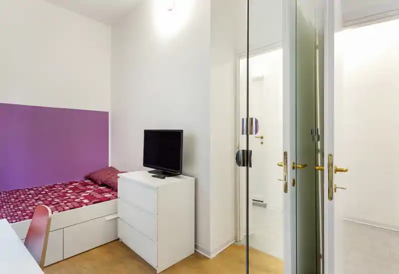 Bright, compact bedroom with a single bed against a purple-accent wall, a white chest of drawers with a TV and a mirrored door opening to a hallway. Clean modern styling with wooden floor and simple textiles.