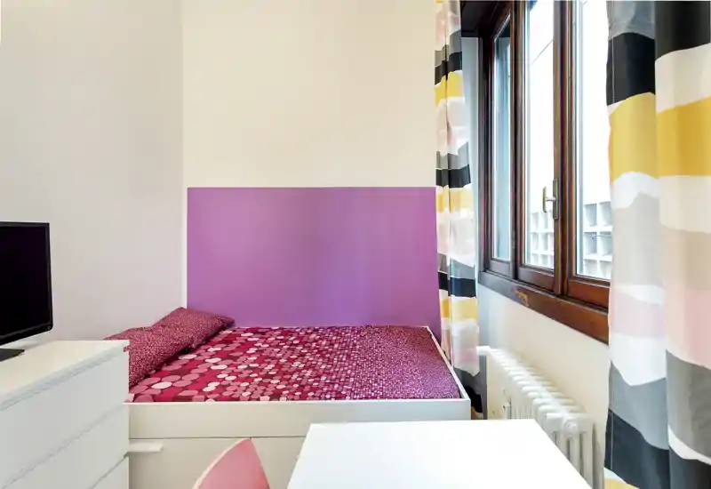 Compact, well-lit bedroom with a colorful double bed, patterned curtains and a wooden-framed window providing natural light — tidy and ready for guests.