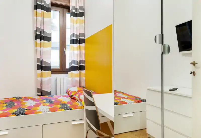 Bright, modern single bedroom with colorful bedding, a study desk and built-in drawers — ideal for a cozy rental. Natural light from the window and a warm yellow accent wall add character.
