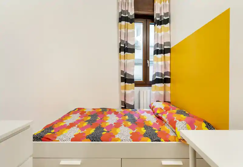 Clean, compact bedroom with a colorful patterned duvet, matching curtains and a yellow accent wall that creates a bright, modern feel.
