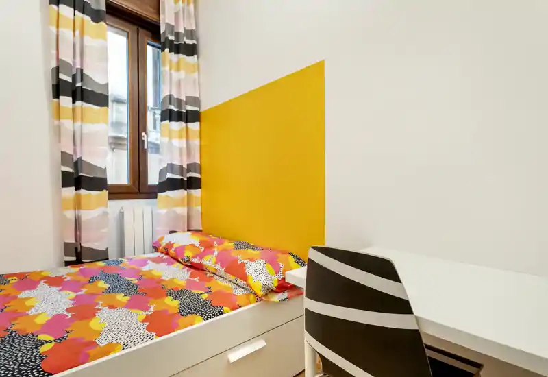 Small, well-lit bedroom with a single bed dressed in colorful patterned bedding, a yellow headboard accent and a white desk with chair — ideal for a cozy single stay.