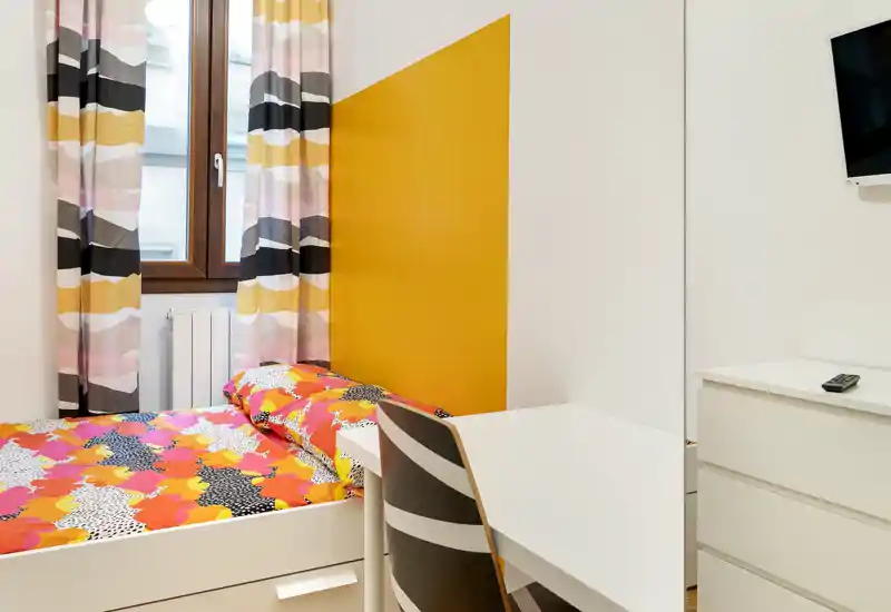 Compact, colorful bedroom with a single bed, patterned curtains and a small desk — bright and freshly styled for a comfortable stay.