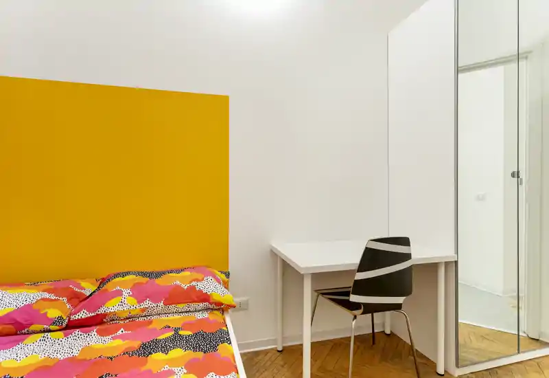 Bright, modern bedroom with a bold yellow headboard and colorful patterned bedding; small white desk and mirrored wardrobe complete the setup.