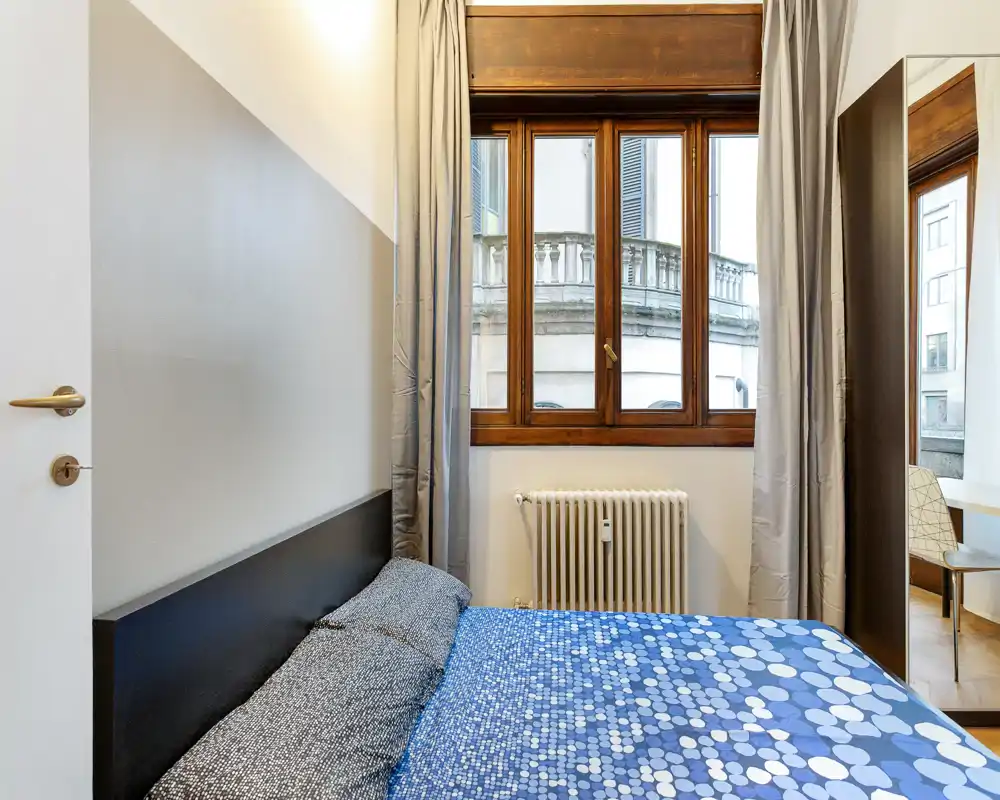 Compact bedroom with a patterned double bed positioned under a large wooden-framed window; neutral walls and light curtains create a calm, well-lit space.