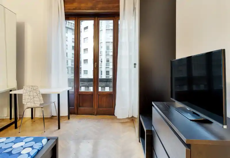 Camera da letto luminosa e arredata in modo semplice con porta finestra in legno verso il balcone, un letto singolo nell'angolo, una scrivania compatta con sedia e una TV su un comò scuro.