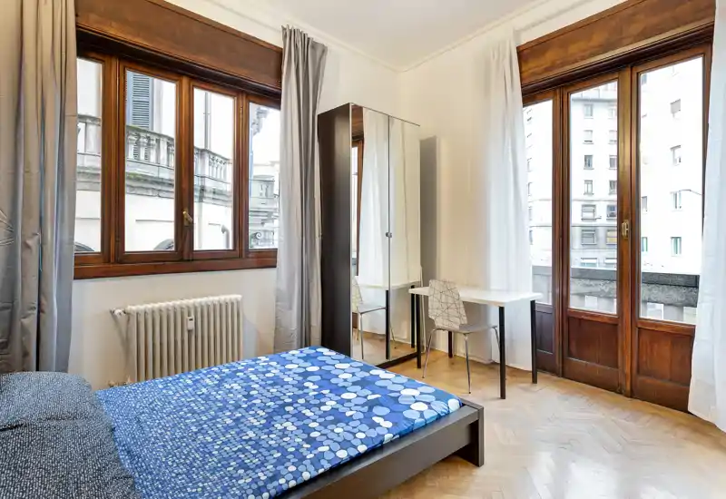 Camera da letto luminosa e arredata in modo essenziale con grandi finestre e un letto matrimoniale con copriletto blu; parquet e un armadio con specchio rendono l'ambiente spazioso.