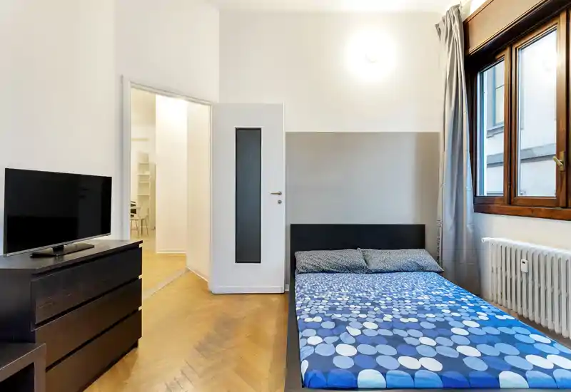 Camera da letto luminosa e ordinata con letto matrimoniale su parete neutra, pavimento in legno, ampia finestra e comò con TV — spazio semplice e ben illuminato ideale per soggiorni brevi.