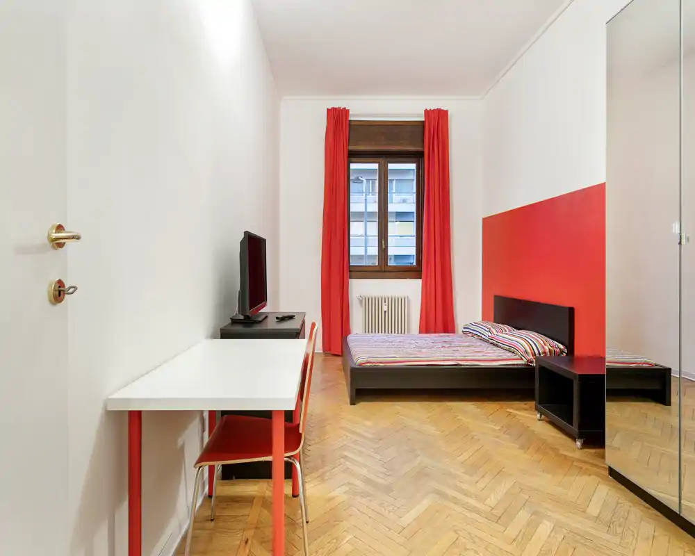 Bright, modern bedroom with a low platform bed, colorful bedding and matching red curtains. Wooden herringbone floor and minimalist desk create a clean, well-styled space ideal for short-term rentals.