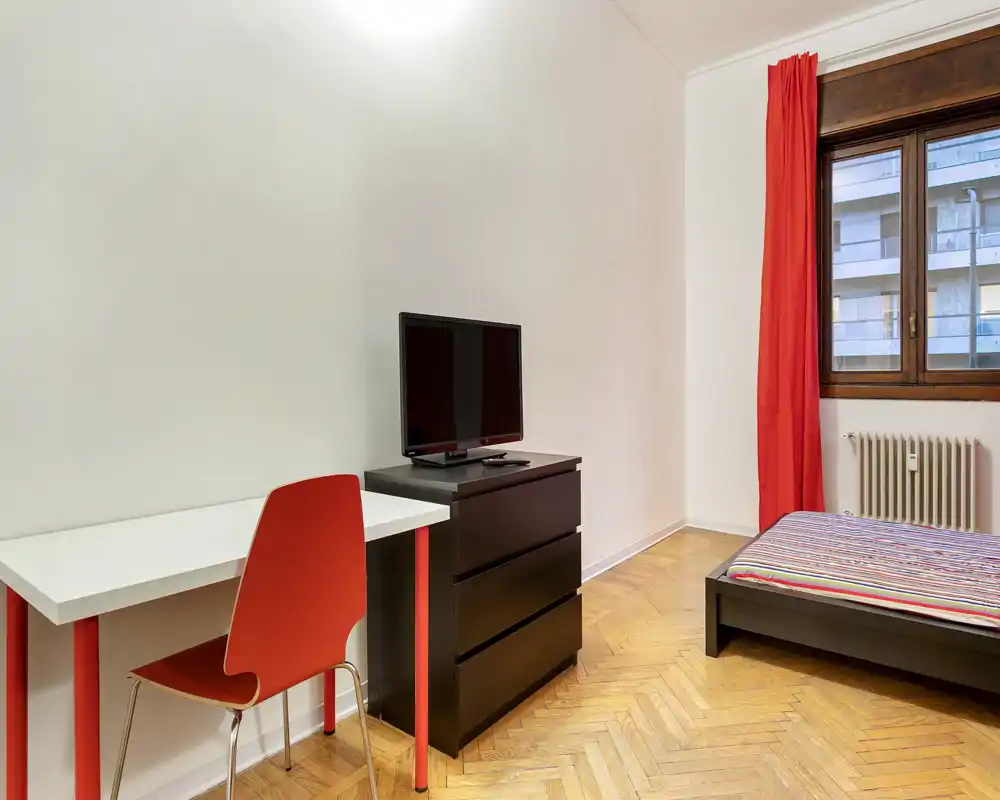 Bright, minimal bedroom with a low platform bed, striped bedding and red curtains; a small desk and chair plus a dresser with TV create a functional, uncluttered space.