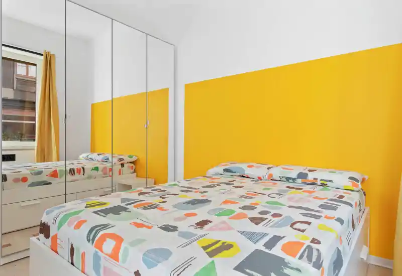 Camera da letto moderna e luminosa con letto matrimoniale con biancheria colorata a motivo, parete accentuata gialla e armadio con ante a specchio che ampliano lo spazio.