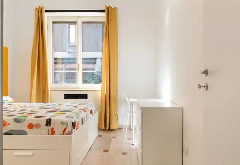 Camera da letto luminosa e moderna con letto contenitore bianco e biancheria colorata vicino a una grande finestra con tende gialle, dotata di scrivania compatta e cassettiera — perfetta per un soggiorno singolo confortevole.