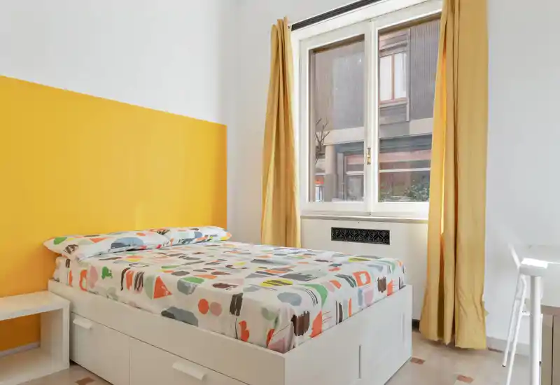 Camera da letto luminosa e moderna con letto colorato, parete accentuata gialla e grande finestra che offre molta luce naturale.