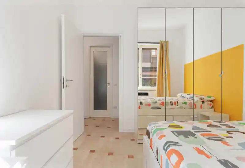 Camera da letto moderna e luminosa con letto colorato, armadio con ante specchiate e parete accentata gialla; la luce naturale rende l'ambiente arioso e accogliente.