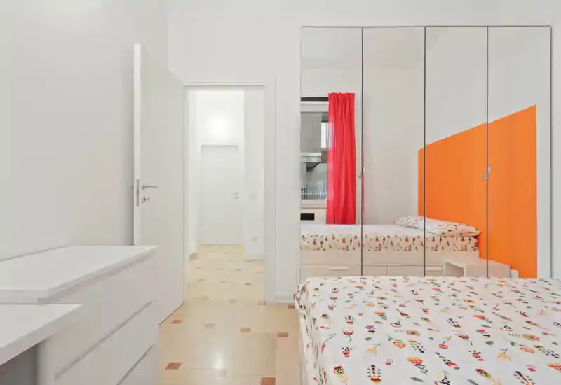 Camera da letto moderna e luminosa con biancheria a fantasia, parete arancione come elemento d'accento e armadio con ante specchiate che ampliano gli spazi; ambiente pulito e accogliente.