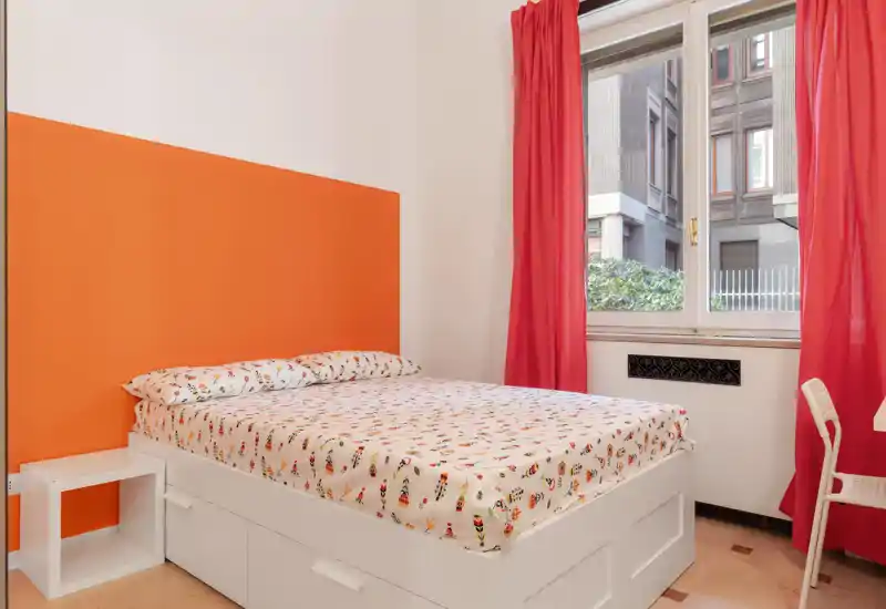 Camera da letto luminosa e compatta con un grande letto bianco con lenzuola a fantasia, testiera arancione e luce naturale dalla finestra con tende rosse.