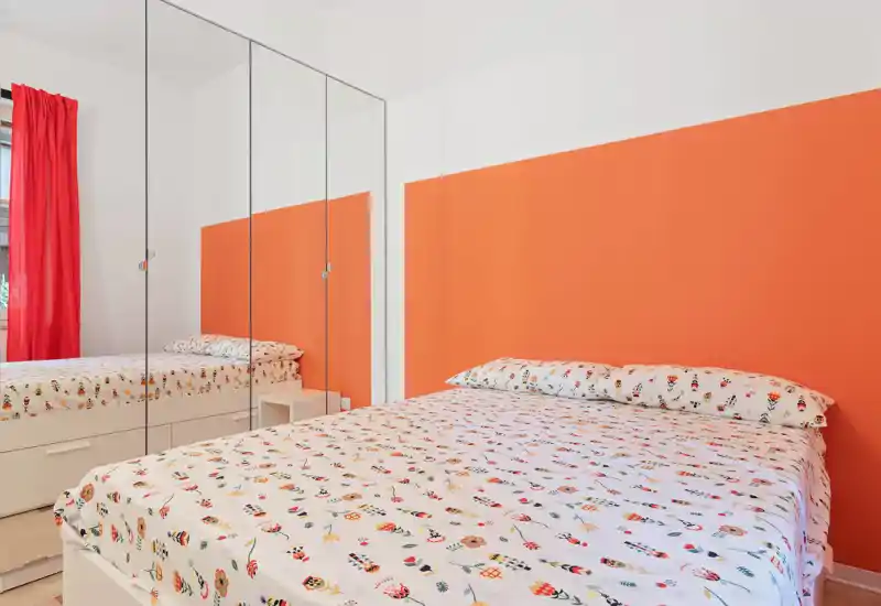 Camera da letto luminosa e ordinata con letto matrimoniale stampato davanti a una parete arancione e armadi con ante a specchio che amplificano la luce.