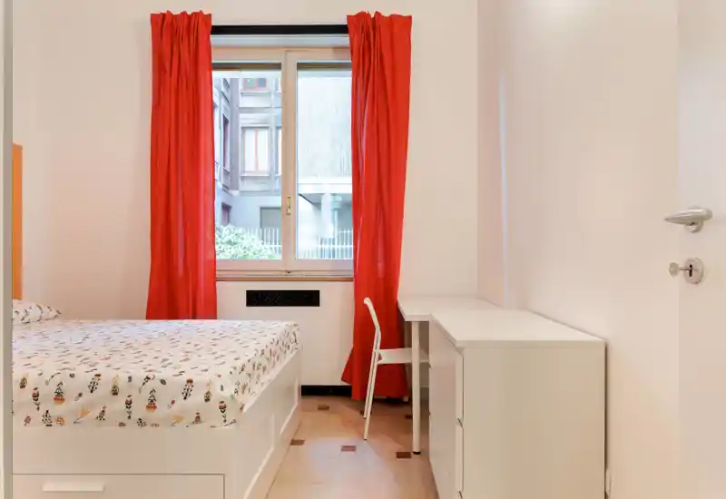 Camera da letto luminosa e ordinata con letto, scrivania e tende rosse vivaci davanti a una grande finestra; arredo minimale e pulito ideale per un annuncio.
