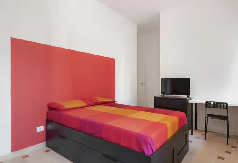 Camera da letto luminosa e moderna con un letto matrimoniale colorato contro una parete rossa, una scrivania e TV — pulita, ben illuminata e minimal.