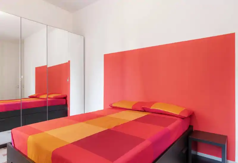 Camera da letto moderna e luminosa con letto matrimoniale con biancheria rosso-arancione, armadio con ante a specchio e comodino minimalista.