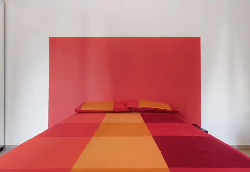 Camera da letto luminosa e minimal con un forte blocco di colore rosso-arancio dietro un letto matrimoniale centrato, con biancheria a quadri coordinata. L'immagine mette in risalto linee pulite, tessuti vivaci e un'estetica moderna e essenziale.