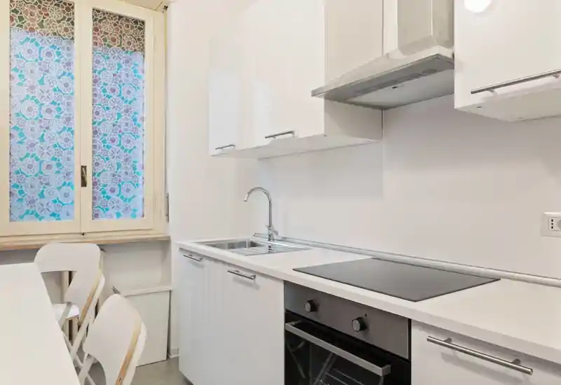 Cucina moderna e luminosa con mobili bianchi, piano cottura integrato e forno, lavello in acciaio e piccolo angolo pranzo accanto a una finestra decorativa.