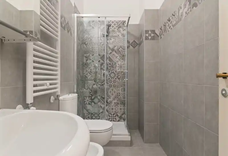 Bagno moderno e compatto con doccia a vetro con piastrelle decorate, radiatore portasciugamani a parete, WC e lavabo bianco su pavimenti e pareti grigie neutre.