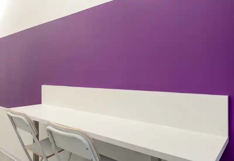 Postazione essenziale con due sedie pieghevoli e un lungo tavolo bianco contro una parete viola — area pulita e semplice ideale per lavoro o studio brevi.