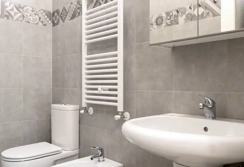 Bagno moderno e compatto con lavabo in ceramica bianca, WC e bidet, piastrelle grigie neutre e termoarredo: ambiente pulito e luminoso.