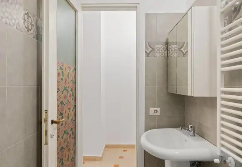 Bagno pulito e compatto con lavabo bianco, mobile con specchio e piastrelle grigie decorative. Spazio luminoso e ben illuminato adatto per un annuncio.