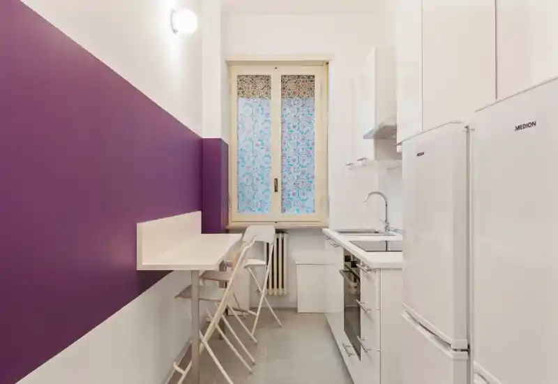 Cucina moderna e compatta con pensili bianchi, forno e frigorifero integrati e un piccolo piano colazione a muro evidenziato da una fascia viola — luminosa e funzionale per la vita in città.