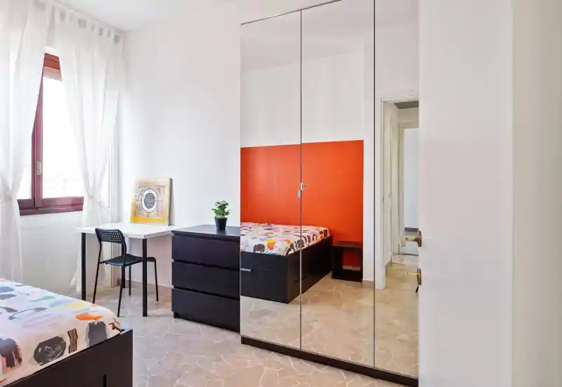 Camera da letto luminosa e moderna con letto matrimoniale davanti a una parete arancione, armadio con ante a specchio, scrivania vicino alla finestra e biancheria colorata — ambiente pulito e arioso, ideale per affitti brevi.