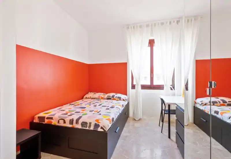 Camera singola luminosa e ben composta con struttura letto scura moderna, biancheria colorata e una finestra ampia che lascia entrare luce naturale. Scrivania semplice e armadio con specchio rendono la stanza pratica e ariosa.