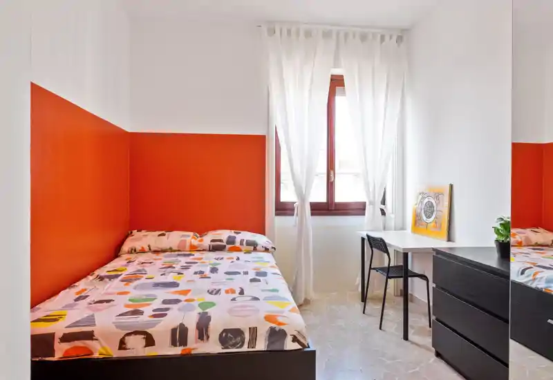 Camera da letto luminosa e accogliente con letto matrimoniale davanti a una parete color arancio, ampia finestra che illumina l'ambiente e una scrivania con comò nero, perfetta per soggiorni brevi in città.