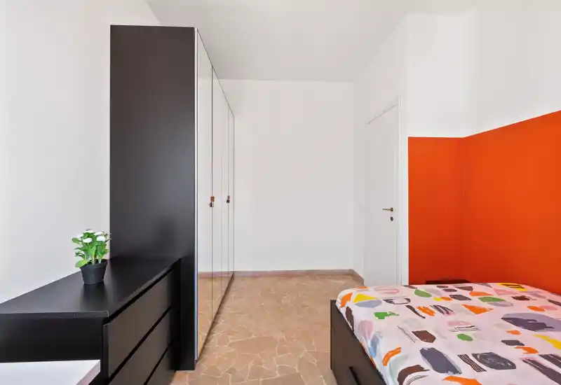 Camera da letto moderna e luminosa con copriletto colorato, armadio con ante a specchio e comò nero dal design essenziale. Linee pulite e una parete arancione a contrasto creano un aspetto fresco e di stile.