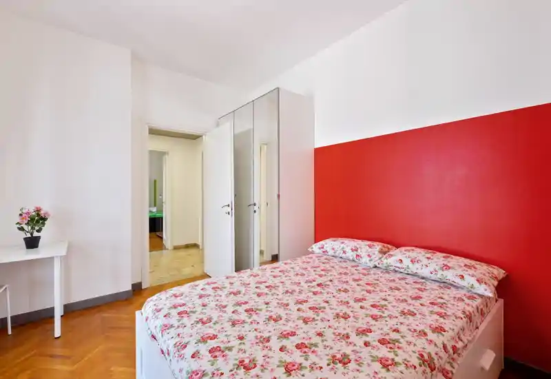 Camera da letto luminosa e ben allestita con un letto matrimoniale coperto da biancheria floreale e una parete rossa a contrasto; mobili bianchi e parquet creano un ambiente pulito e accogliente.