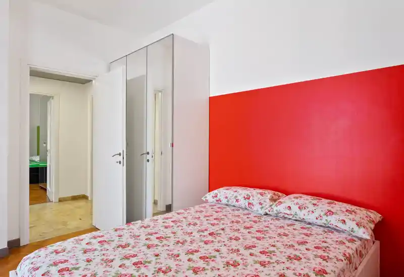 Camera da letto luminosa e ordinata con letto matrimoniale a fiori contro una parete rossa intensa e un armadio con ante a specchio che amplifica la luce.