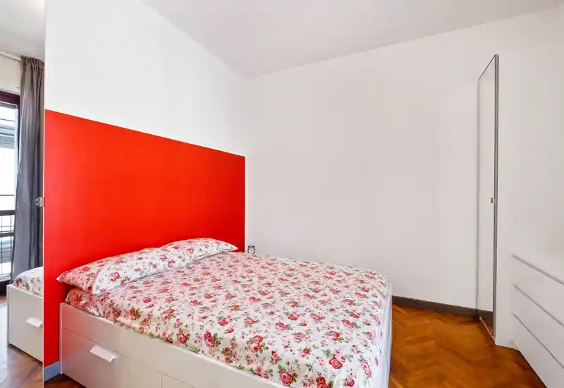 Camera da letto luminosa e ordinata con letto matrimoniale a fiori, testiera rossa a contrasto e pavimento in parquet. La luce naturale valorizza l’arredo essenziale.