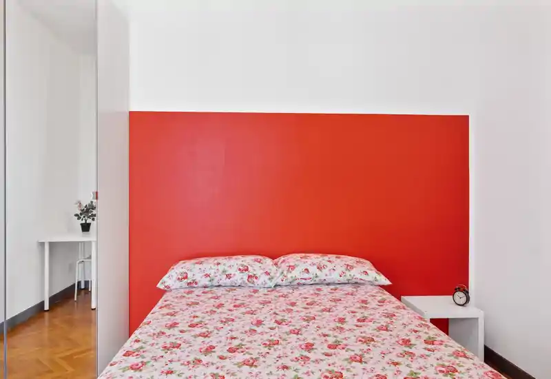 Camera da letto luminosa e ordinata con letto matrimoniale con biancheria floreale su uno sfondo con parete rossa; comodino semplice con sveglia e una piccola scrivania visibile vicino all'armadio con specchio.