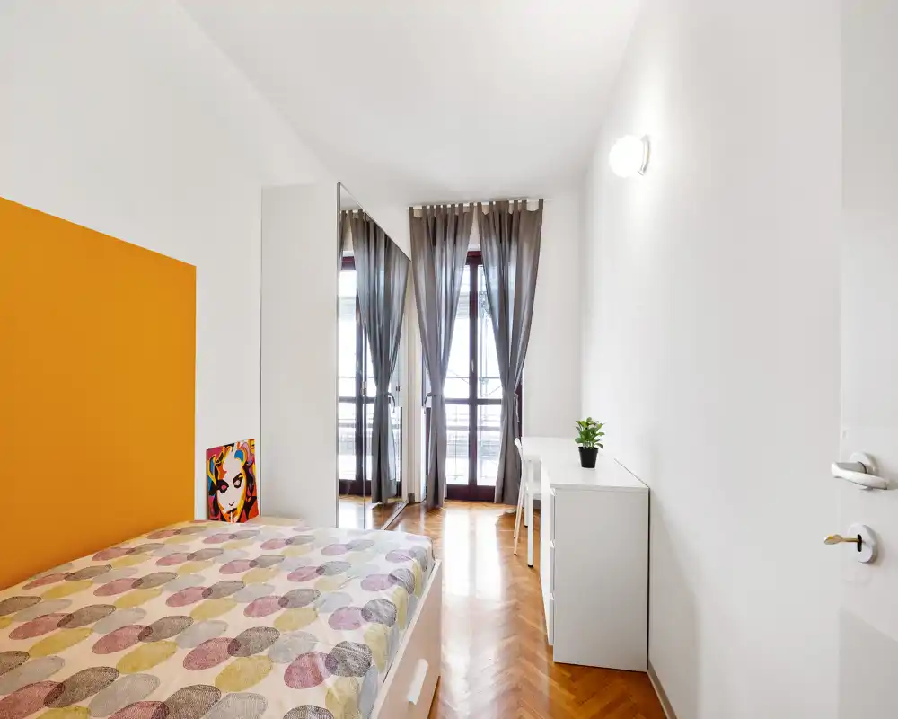 Camera da letto moderna e luminosa con letto matrimoniale con copriletto fantasia, pavimento in parquet a spina di pesce e porte sul balcone con tende leggere — mobili bianchi e un tocco di colore sulla parete.