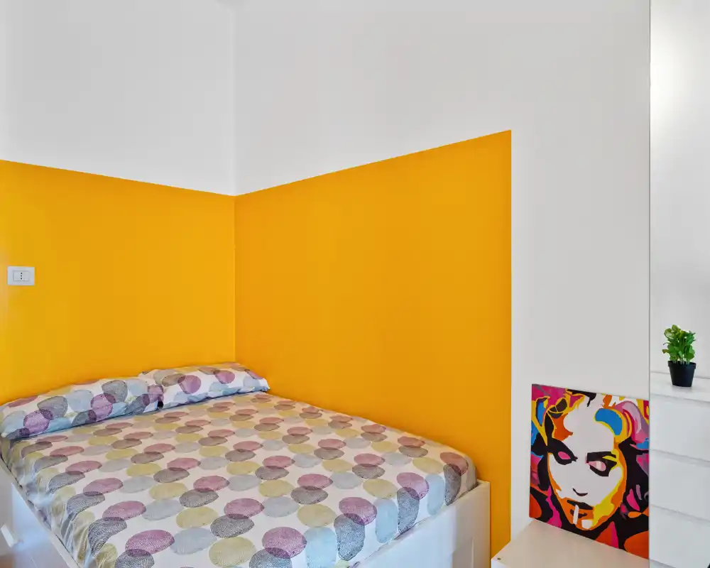 Camera da letto luminosa e moderna con letto matrimoniale davanti a una parete gialla, biancheria a motivi e pochi elementi decorativi che la rendono accogliente e ben illuminata.