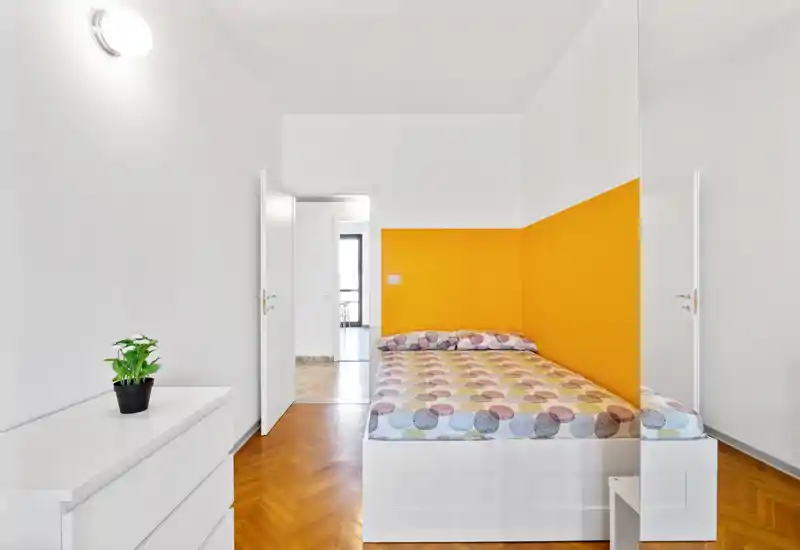 Dormitorio moderno y luminoso con cama doble junto a una pared amarilla, ropa de cama estampada y muebles blancos minimalistas; suelo de madera pulida y luz natural crean un ambiente acogedor.