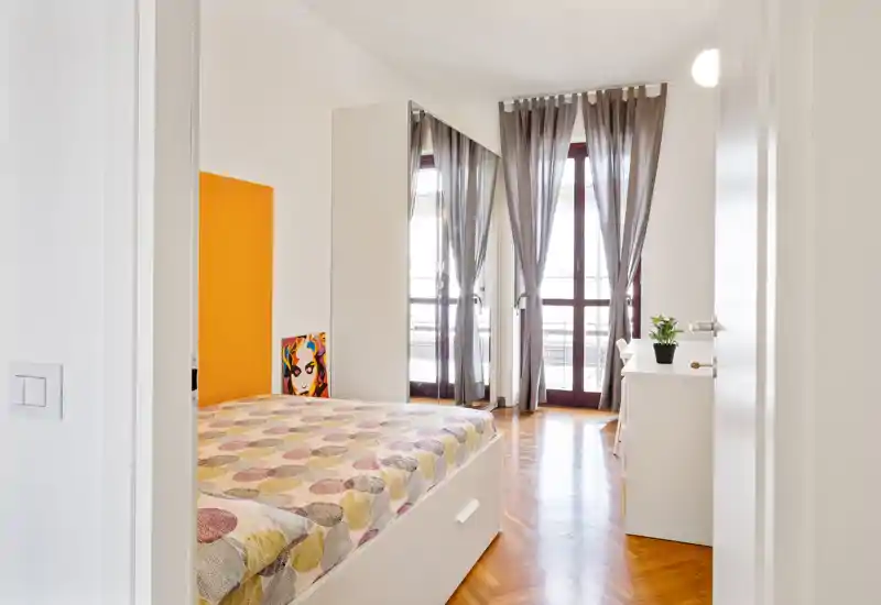 Dormitorio luminoso y moderno con cama doble de estampado, suelo de parquet y grandes puertas al balcón; mobiliario blanco y cortinas ligeras para un ambiente limpio.