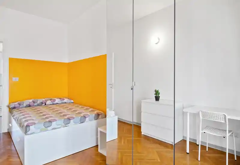 Dormitorio moderno y luminoso con cama completa junto a una pared amarilla, armario con espejo, mobiliario blanco y un pequeño escritorio — espacio limpio y acogedor, ideal para una persona o pareja.