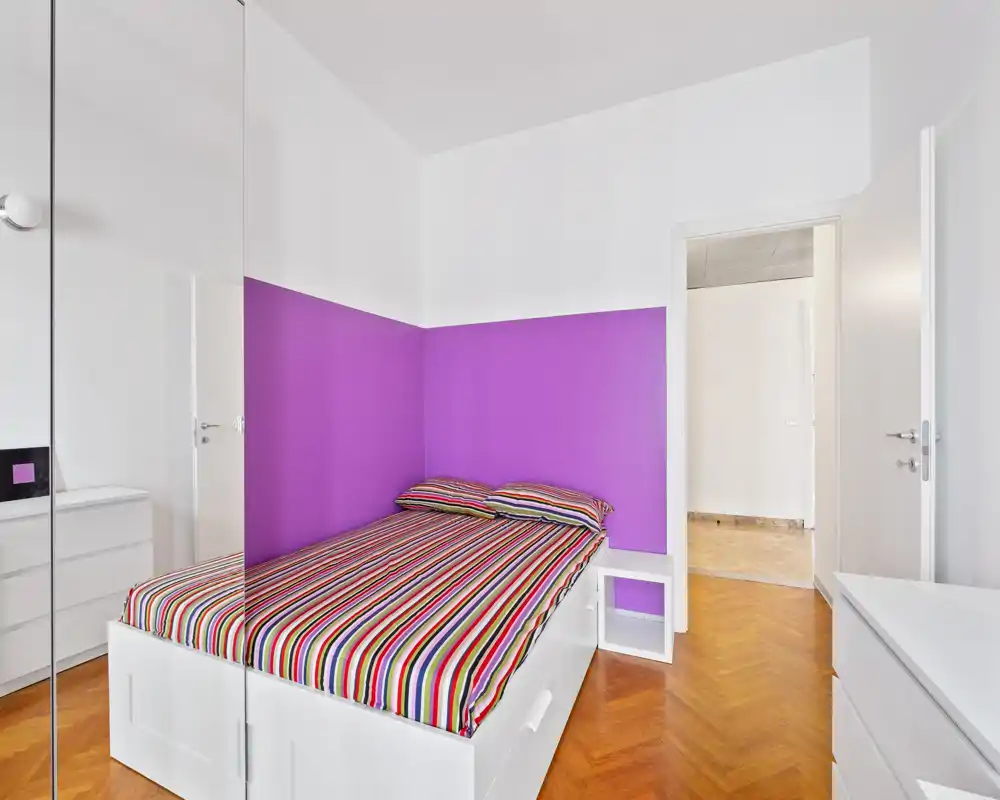 Dormitorio moderno y luminoso con cama blanca frente a una pared morada, ropa de cama a rayas y mobiliario blanco — bien iluminado y preparado para su publicación.