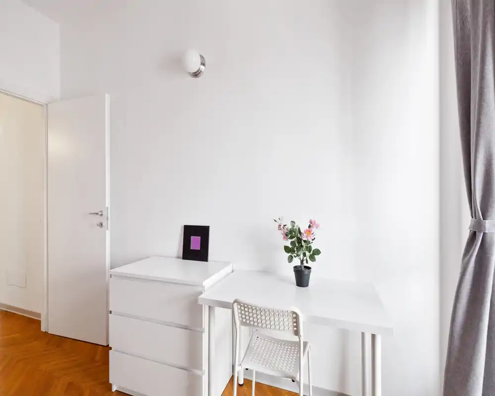 Rincón de salón minimalista y luminoso con mesa blanca, cómoda y silla; una planta en maceta y un marco añaden estilo y luz natural desde la ventana con cortina.